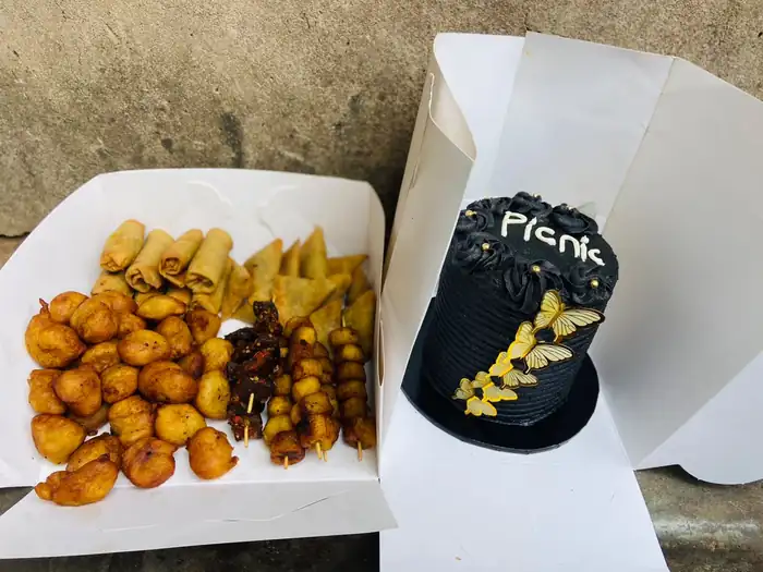 Small chops box with puff-puff, spring rolls and peppered meat beside a black and gold "Picnic" cake